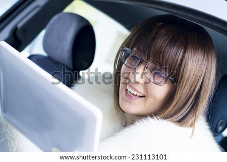 Beautiful woman in white fur coat smiling while using computer in a car