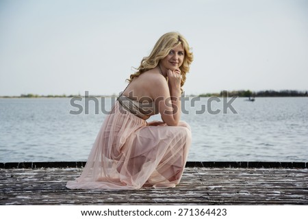 Beautiful blond curly woman wearing evening peach color gown squatted on the deck at the lake. Fashionable and glamorous dress and style.