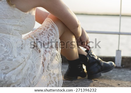 Back of a blond woman wearing white lace dress and short black boots looking to the lake enjoying sun. Fashion for young people.