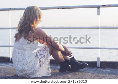 Back of a blond woman wearing white lace dress and short black boots looking to the lake enjoying sun. Fashion for young people.