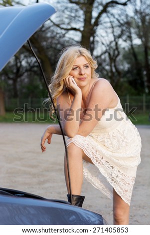 Blond woman wearing white lace dress and short black boots in front of the trunk of a car thinking what to do after car break down.