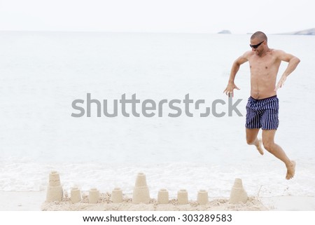 Man smiling and wearing sun glasses and blue shorts jumping above sand castle on beach wanting to destroy it. Beach fun.