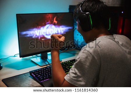 Portrait of happy teenage gamer boy winning while playing video games on computer in dark room wearing headphones and using backlit colorful keyboard