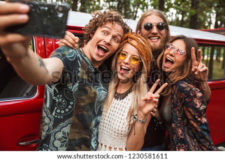 Group of young hippy men and women smiling and taking selfie on mobile phone near vintage minivan into the nature