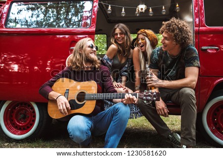 Group of excited hippies men and women having fun and playing guitar near vintage minivan into the nature