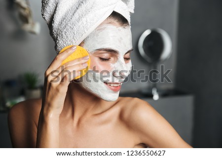 Beautiful young woman with towel wrapped around her head removing face mask with a sponge at the bathroom