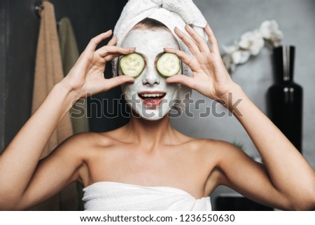 Beautiful young woman with towel wrapped around her head wearing face mask, holding cucumber slices at the bathroom