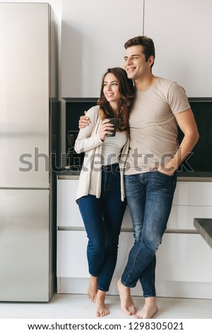 Happy lovely young couple holding cups while standing at the kitchen at home, looking at the window