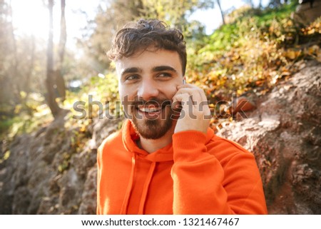 Picture of handsome happy young sports fitness man runner outdoors in park talking by mobile phone.