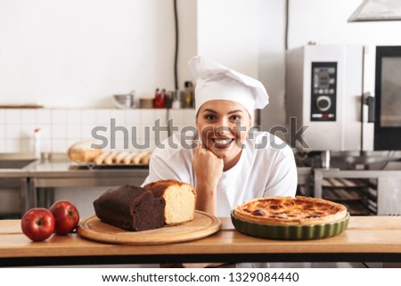 Image of positive woman chef wearing white uniform posing in kitchen at the cafe with baked goods