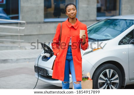 Young african woman is standing near the electric car and looking at camera. The rental car is charging at the charging station for electric vehicles.