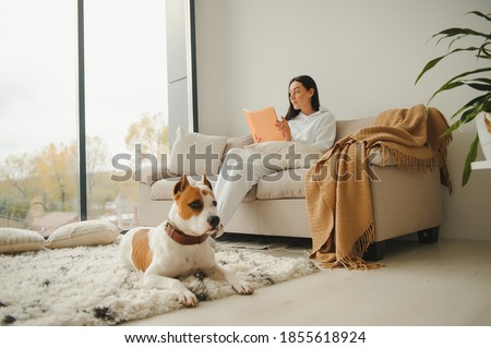 Woman in cozy home clothes relaxing at home with dog reading a book. Comfy lifestyle.