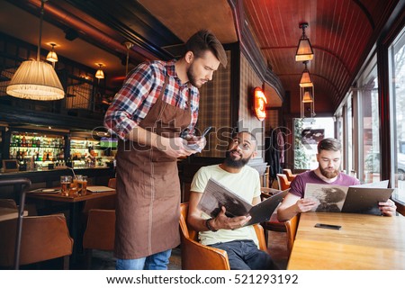 Serious concentrated young waiter standing and taking an order from two bearded handsome men in cafe