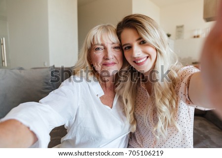 Image of cheerful young lady sitting on sofa at home with her grandmother hugging. Looking camera make selfie.