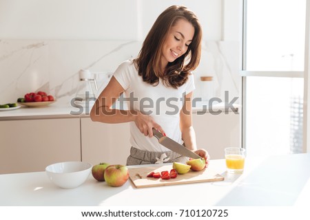 Smiling young woman cutting fruits on a wooden board while making breakfast in a kitchen