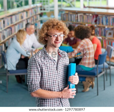 Male high school student in library holding folder smiling at camera