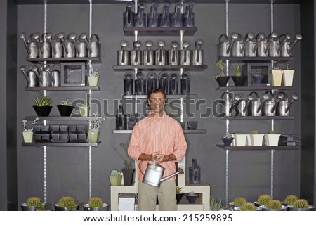 Man standing beside cacti display in shop, holding watering can, smiling, front view, portrait