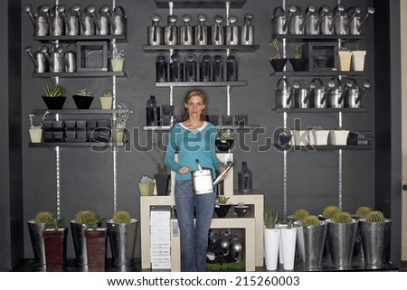 Woman standing beside cacti display in shop, holding watering can, smiling, front view, portrait