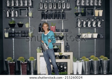 Woman standing beside cacti display in shop, holding watering can, smiling, front view, portrait
