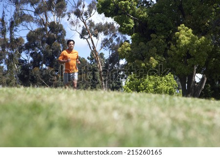 Man wearing orange t-shirt and shorts, jogging on grass in park, surface level, focus on background