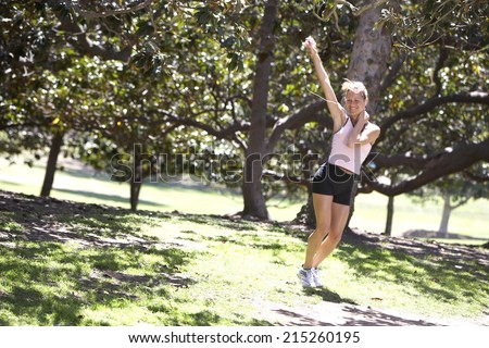 Woman exercising in park, listening to MP3 player, hand raised, smiling, trees in background (tilt)