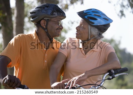 Active senior couple wearing cycling helmets and polo shirts, sitting on bicycles in park, face to face, smiling