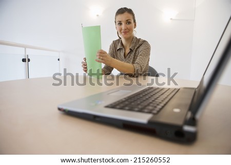 Businesswoman working at desk in office, holding green file, smiling, portrait, laptop in foreground, focus on background, surface level