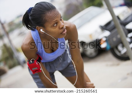 Exhausted female jogger stopping for breath on pavement, leaning on knees, listening to MP3 player strapped to arm, focus on foreground (tilt)