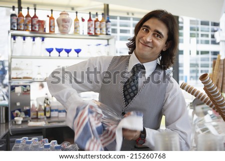 Waiter wiping glass with dishtowel behind bar, smiling, portrait (blurred motion)