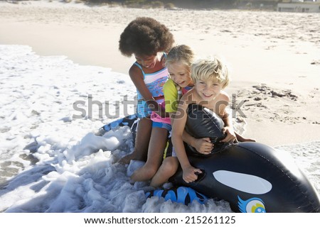 Three children (5-10) sitting on inflatable toy whale on beach, playing in surf, smiling