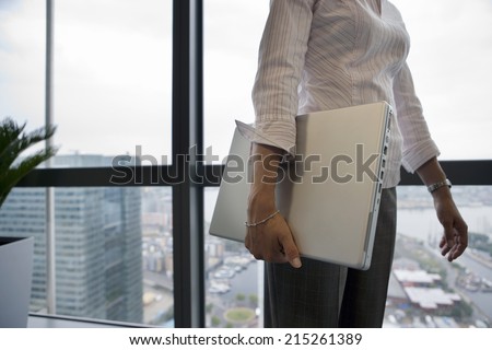 Businesswoman standing beside window, carrying laptop underarm, side view, mid-section