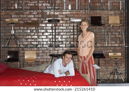 Couple shopping in furniture store, woman standing beside man lying on bed, smiling, portrait