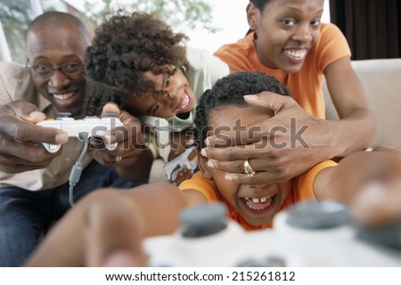 Family playing with video games console at home, mother covering son's (7-10) eyes, smiling
