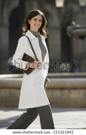 Woman in polo neck jumper and white coat walking in plaza, carrying handbag, smiling, portrait