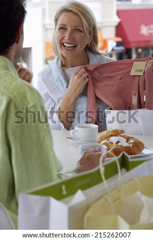 Couple sitting at cafe table, blonde woman showing man new top, laughing