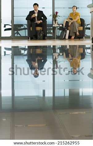 Businessman and businesswoman sitting in lobby, man holding cup, reflection on shiny floor