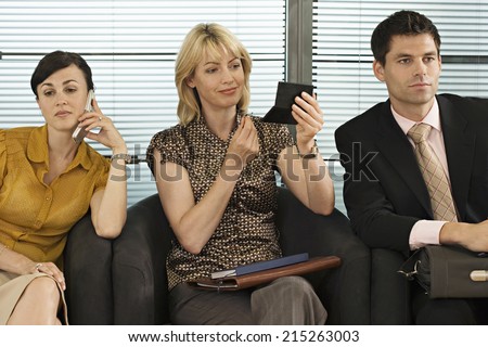 Business people waiting in office reception, woman using mobile phone, second woman applying make-up