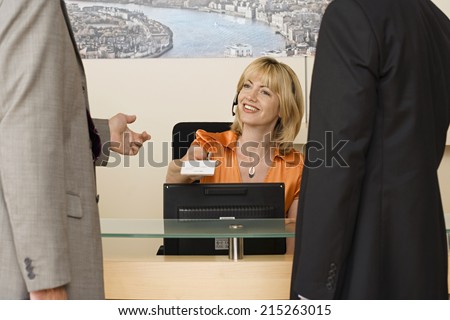 Receptionist working behind reception desk, receiving business card from businessman, smiling