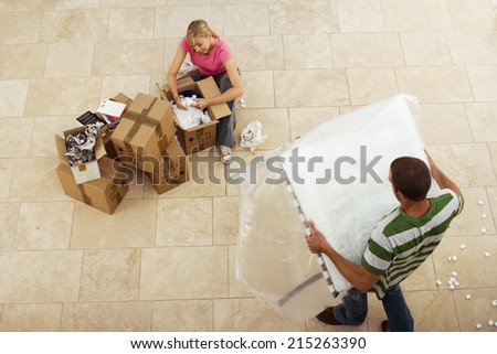 Couple moving house, woman packing crockery in boxes, man carrying table, overhead view
