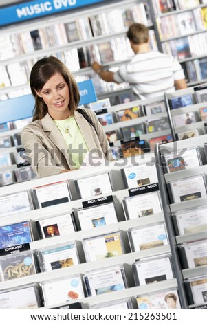 Young woman shopping for CDs in record shop, standing in aisle behind rack, smiling, man in background (differential focus, tilt)