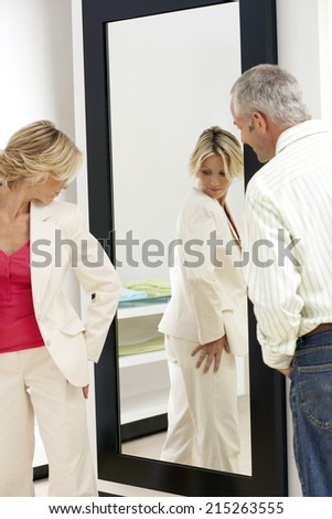 Woman trying on new clothes in fitting room, looking at reflection in mirror, husband watching