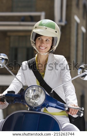 Woman in crash helmet riding on scooter in street, making sideways glance, smiling, front view