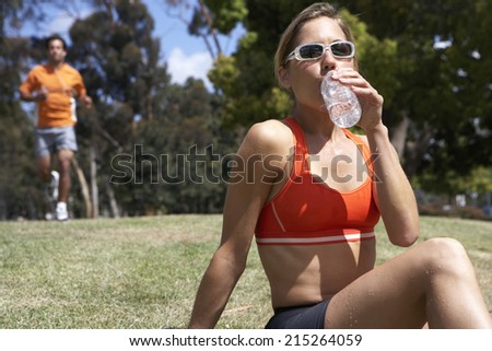 Woman sitting on grass in park, drinking from water bottle, man jogging, focus on foreground