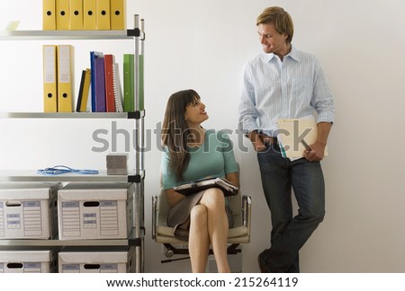 Businessman flirting with businesswoman in office, woman sitting in chair beside shelf, smiling