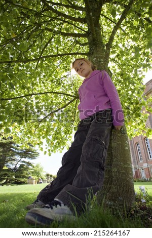 Girl wearing purple top and combat trousers, leaning against tree trunk in garden, head cocked, portrait, low angle view