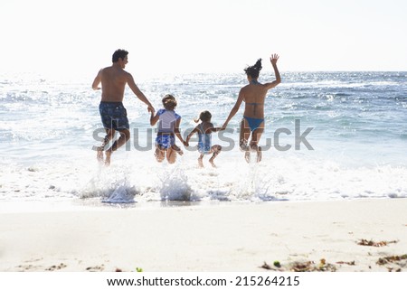 Two generation family wearing swimwear, jumping above surf on sandy beach, side by side, holding hands, rear view, sunlight shimmering on sea
