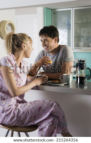 Couple in pyjamas sitting at breakfast bar in kitchen, man holding glass, woman on stool, smiling