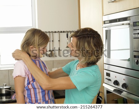Mother and daughter (6-8) standing in kitchen, girl wearing striped apron, woman tying knot, smiling