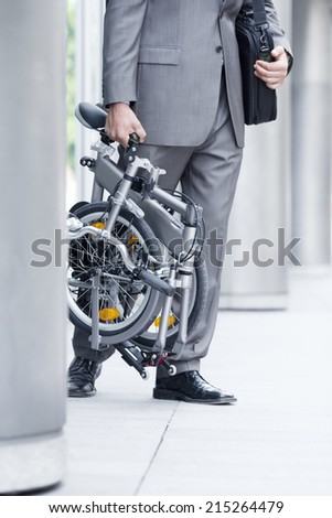 Businessman leaving office building, carrying folding commuter bicycle, side view, low section