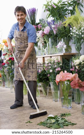 Male florist in apron standing in flower shop, sweeping floor with broom, smiling, portrait